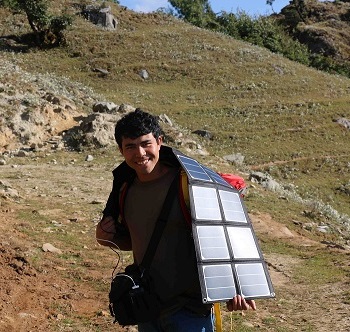 Man carrying a large solar panel on a hillside under sunlight.