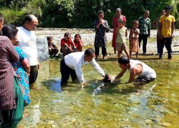 A group of people witness a baptism in a shallow, clear river outdoors.