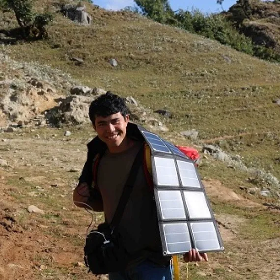 Man carrying a large solar panel on a hillside under sunlight.