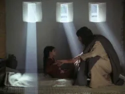 A man comforts a child sitting on a bed with sunlight streaming through three windows.